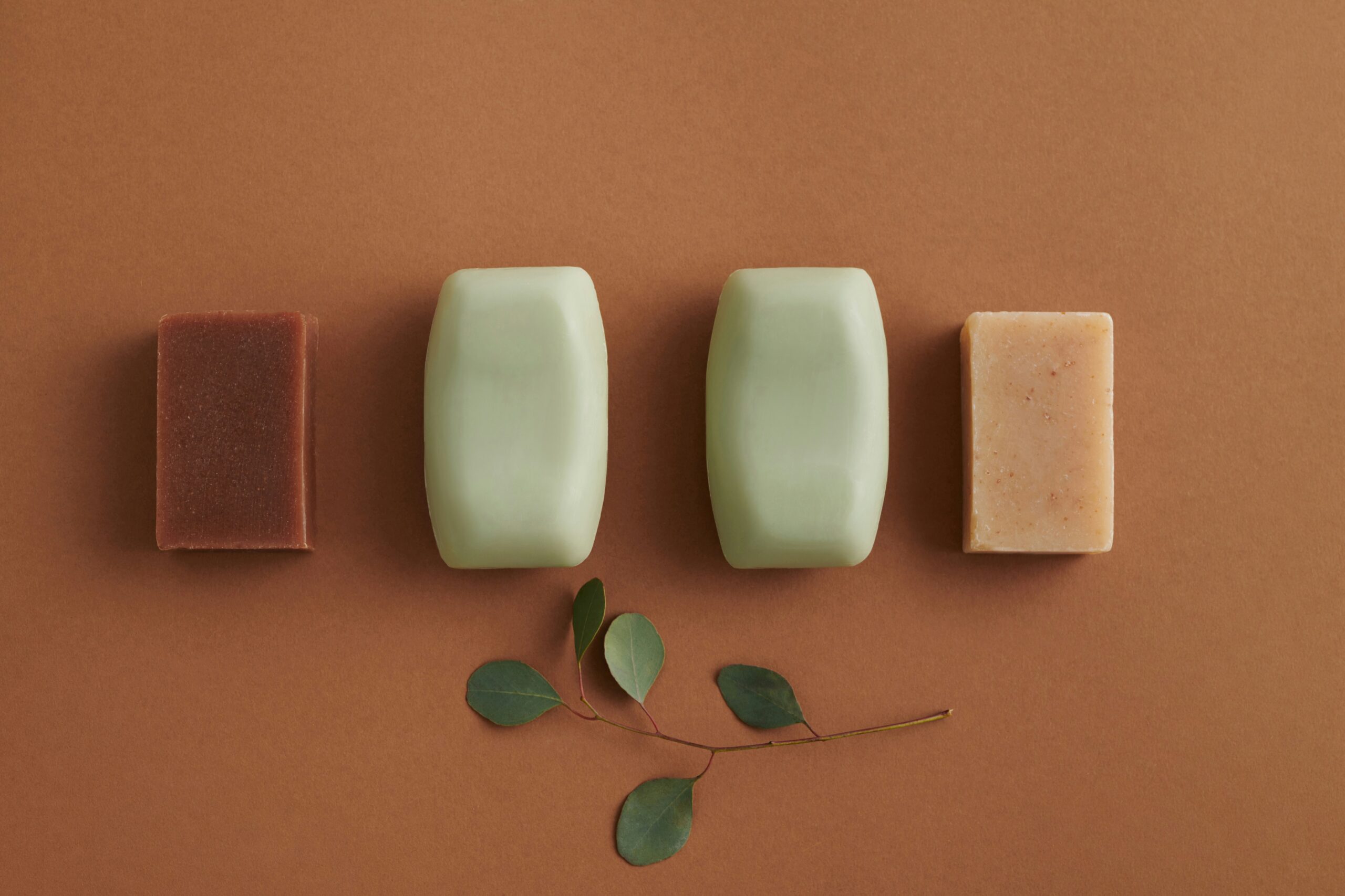 Bar Soap Product Variety – Natural and Certified Soap Manufacturing Four bar soaps in different colors and shapes displayed on a neutral background, with natural green leaves as accent.
