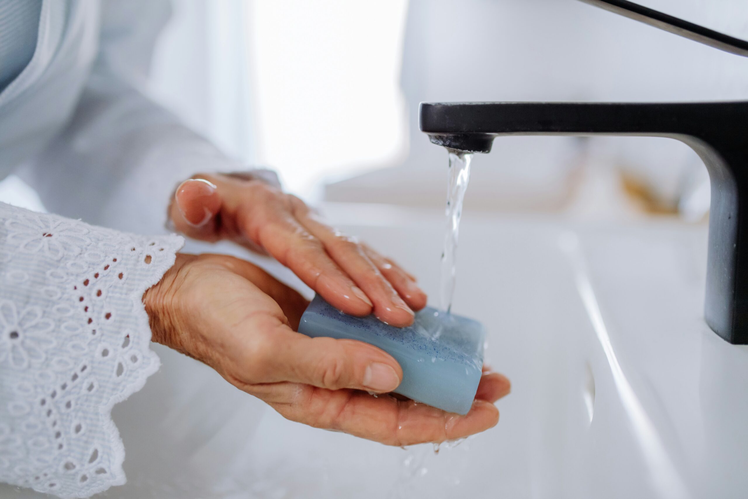 Privacy Policy – Vanguard Soap Manufacturing Hands using a blue bar of soap under running water at a sink, demonstrating lather and cleansing in action.